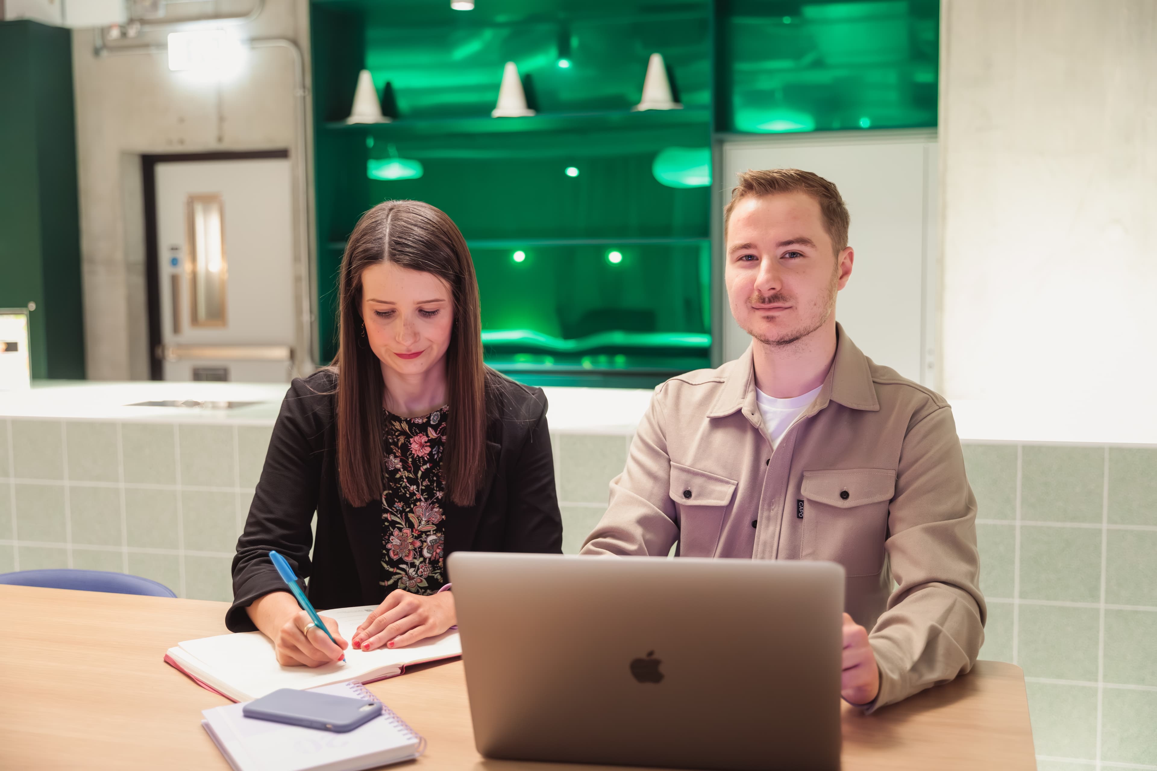 Woman writing in a notebook beside a man with a laptop in a modern office.