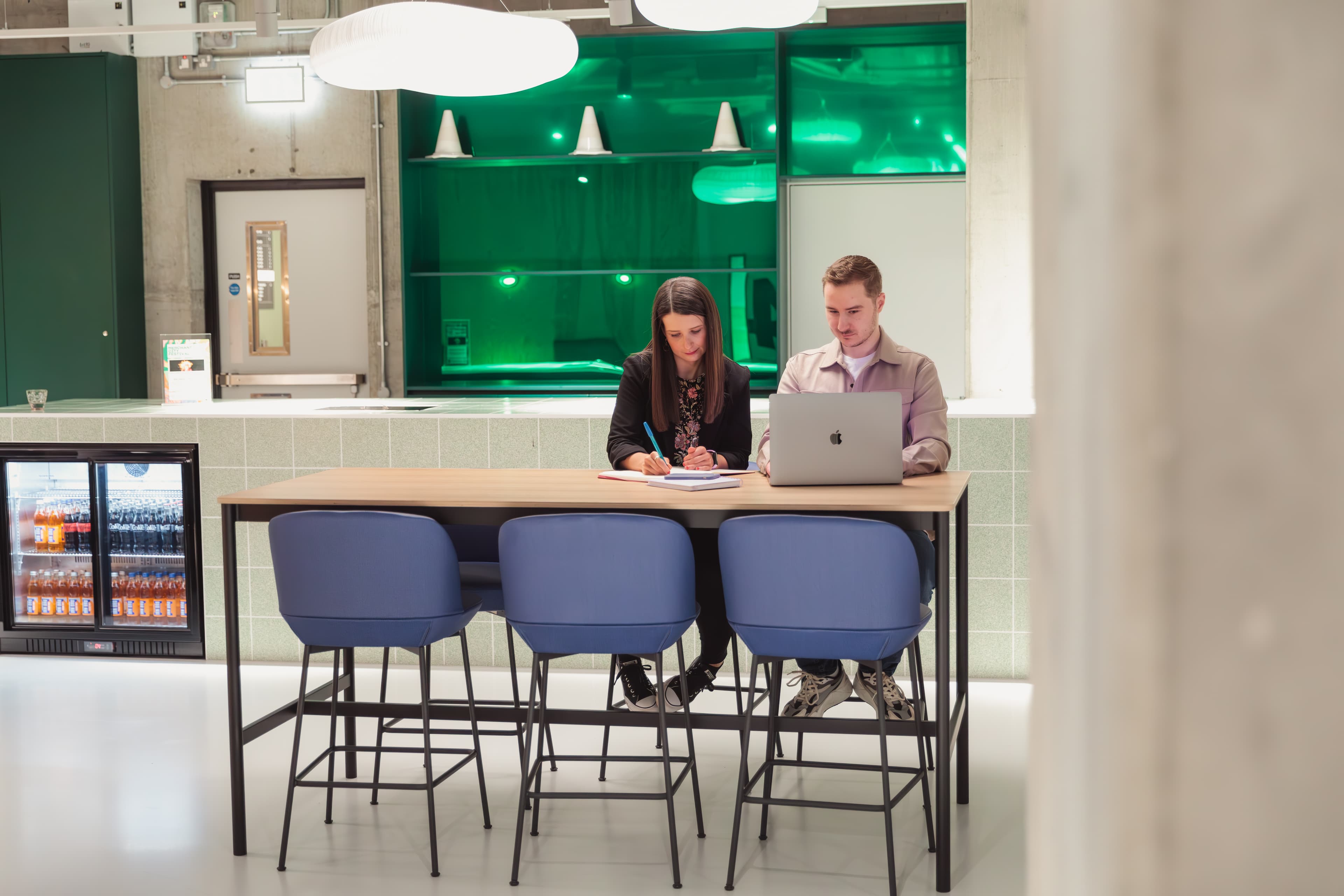 Woman writing and man using a laptop at a high table with blue chairs.