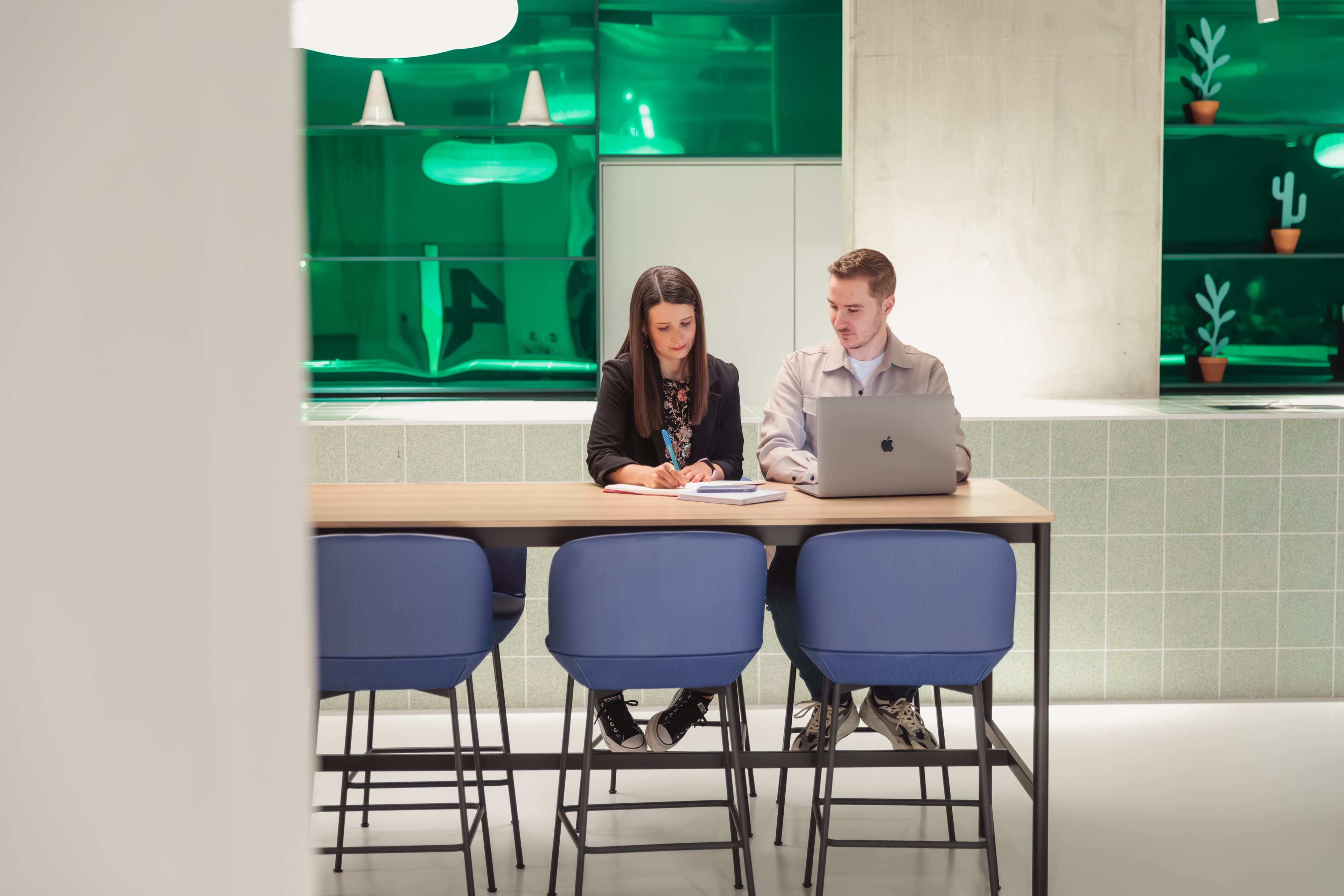 Two colleagues collaborating at a long wooden table with a laptop and notebook in a modern office.