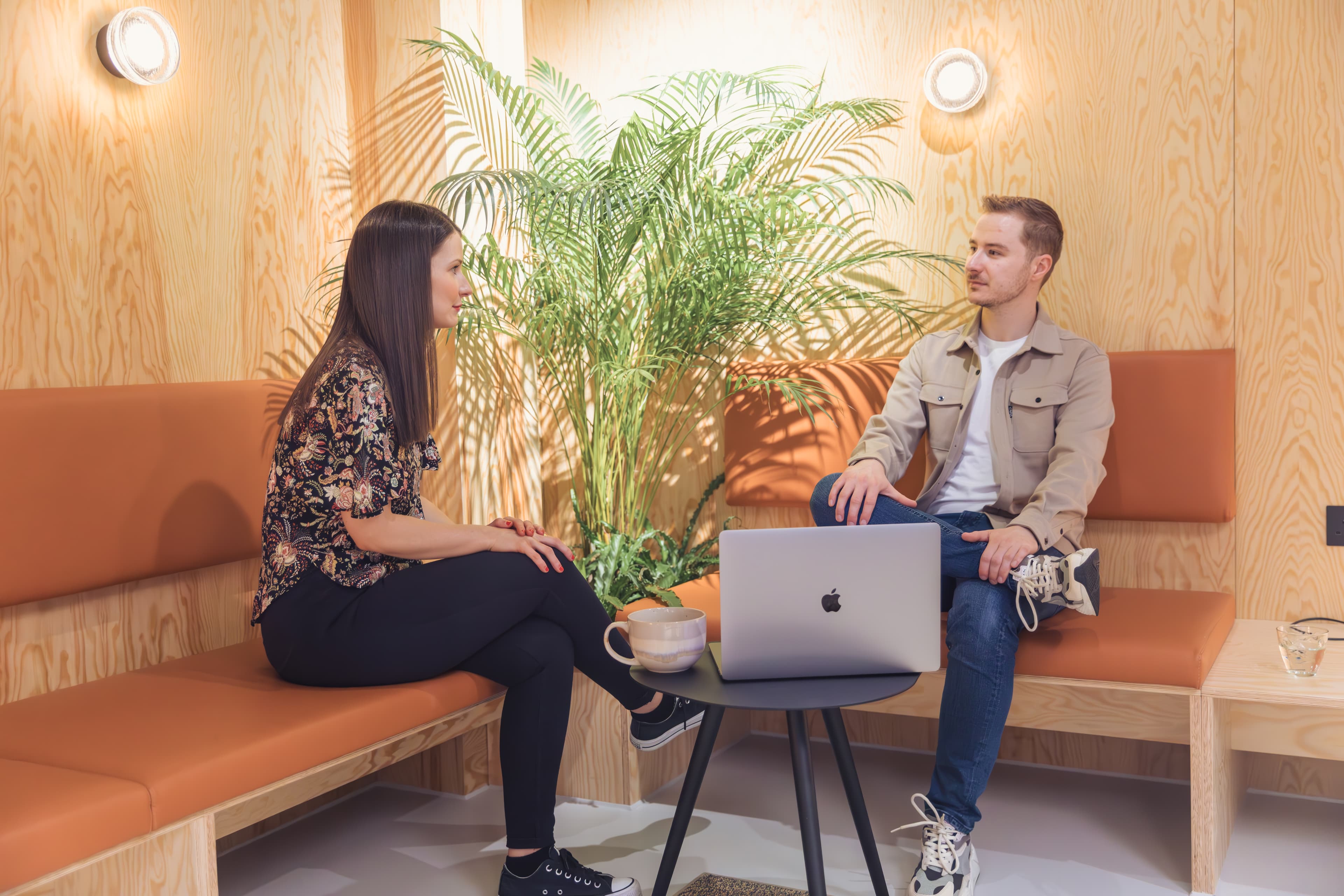 Man and woman talking on an orange bench in a wood-paneled room with laptop.