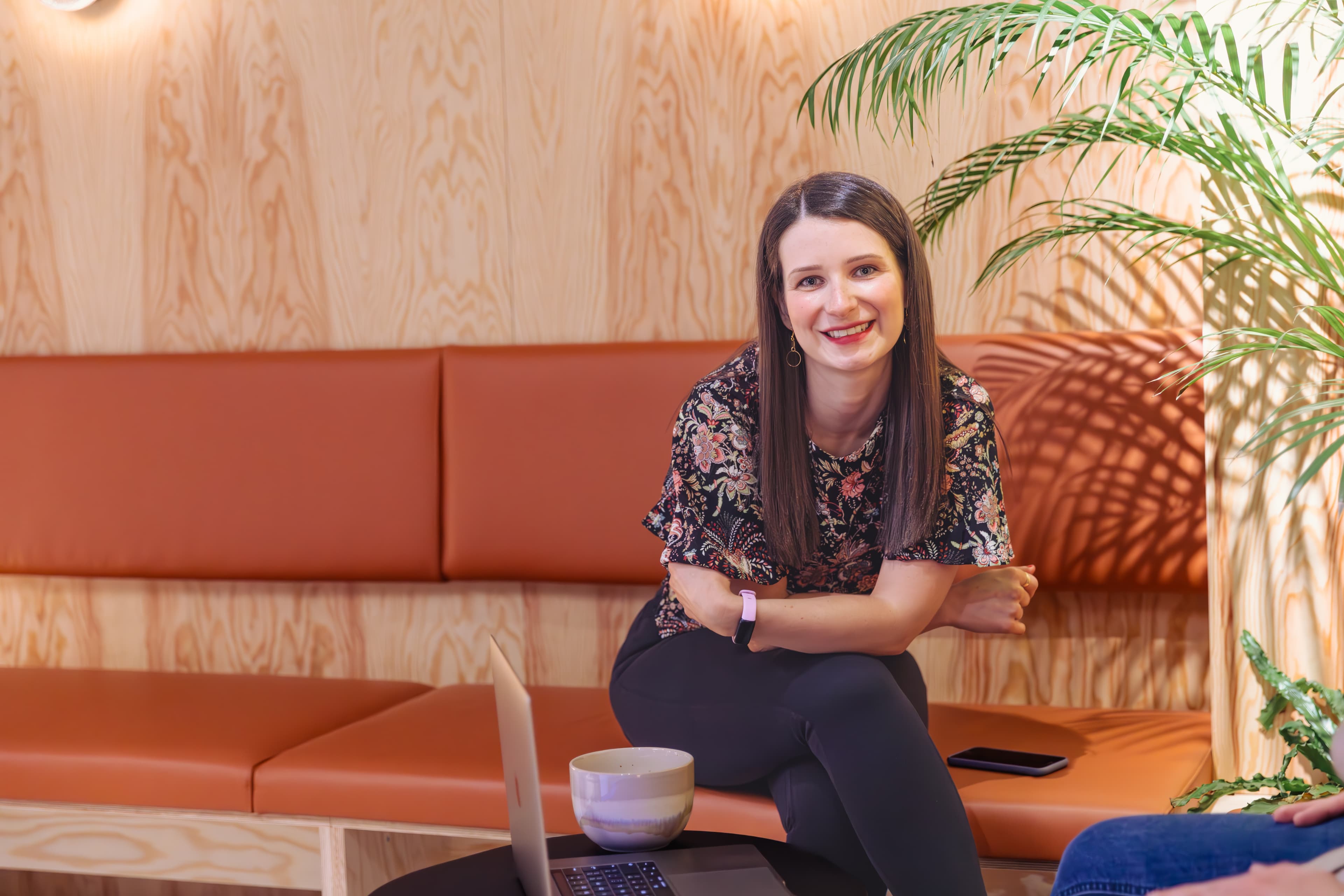 Smiling woman in floral top sits on a brown bench with a laptop nearby.