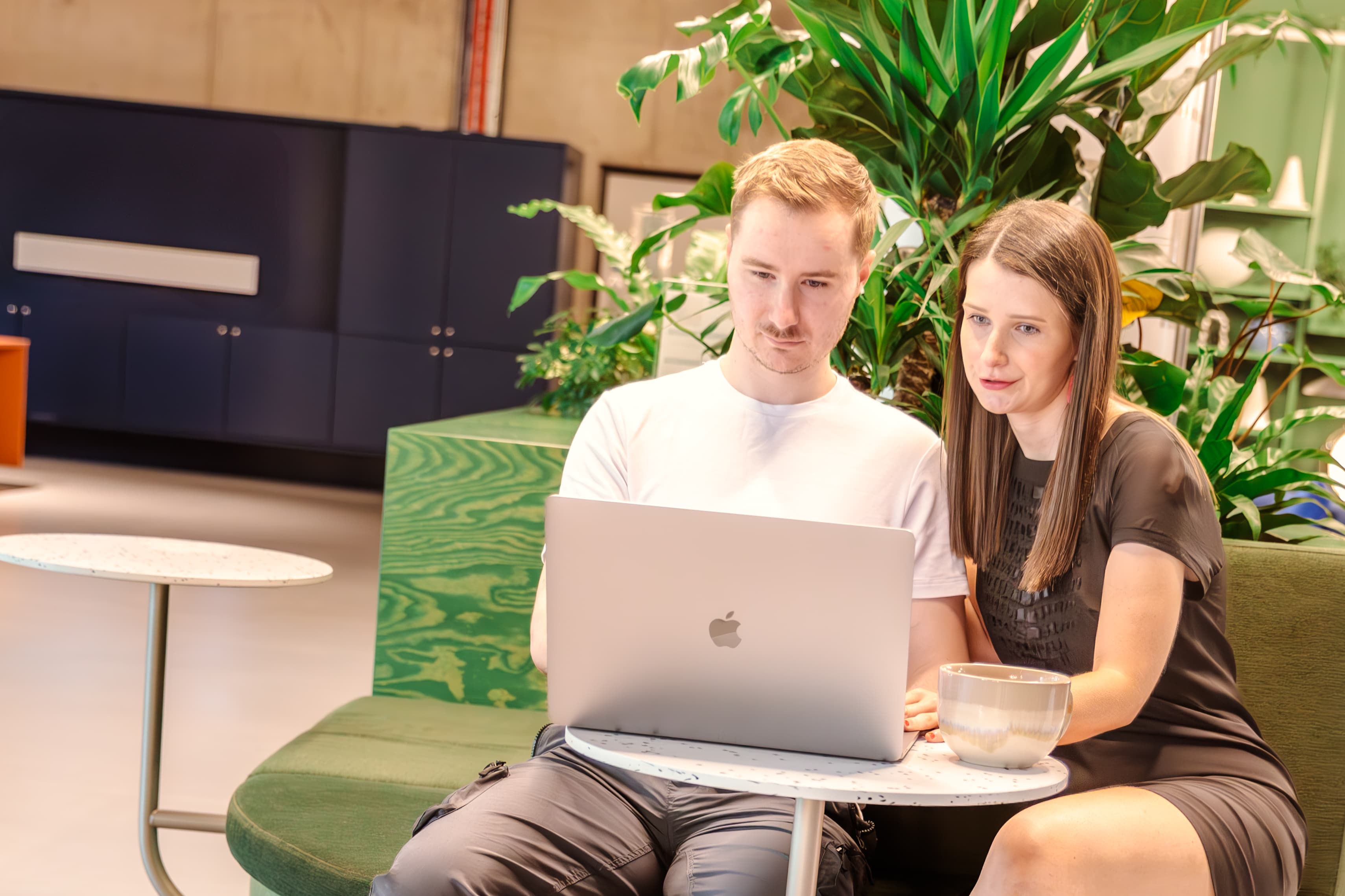 Man and woman sitting on a green bench, looking at a laptop in a leafy office.