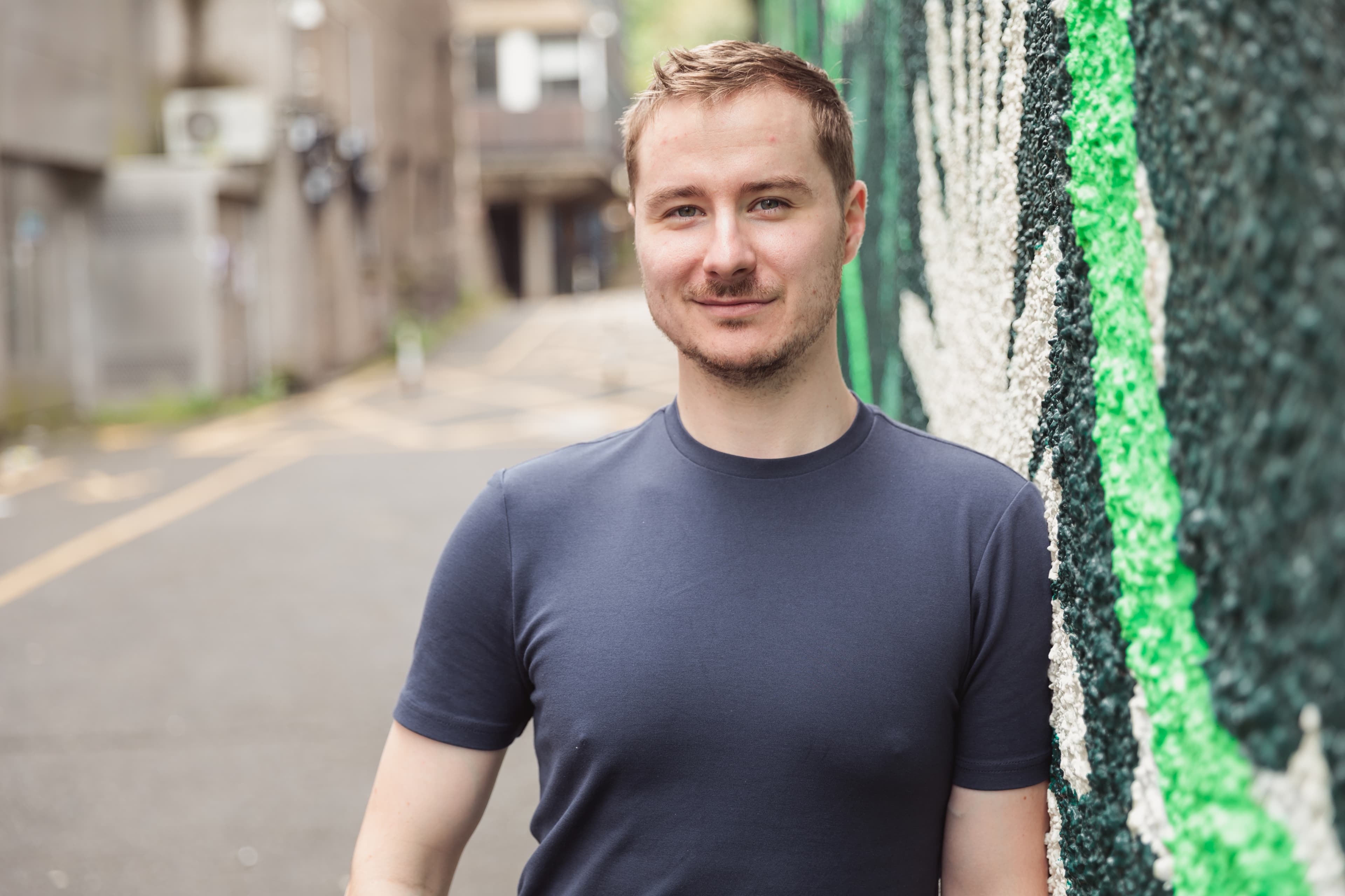 Smiling man in a navy blue t-shirt leaning against a green and white textured wall.