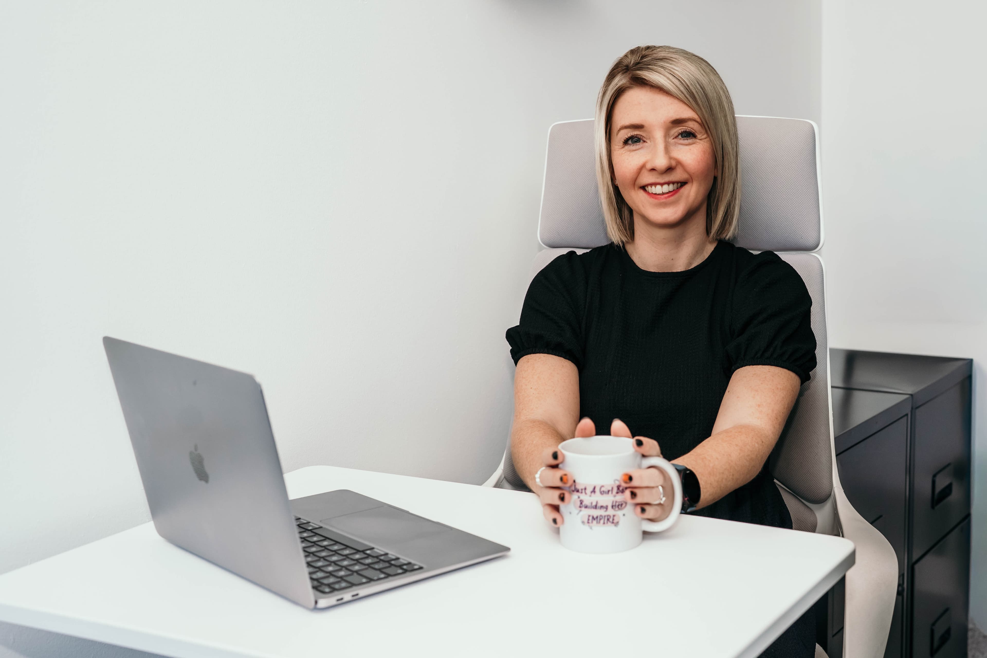 Smiling blonde woman sitting at a white desk with a laptop, holding a white mug.