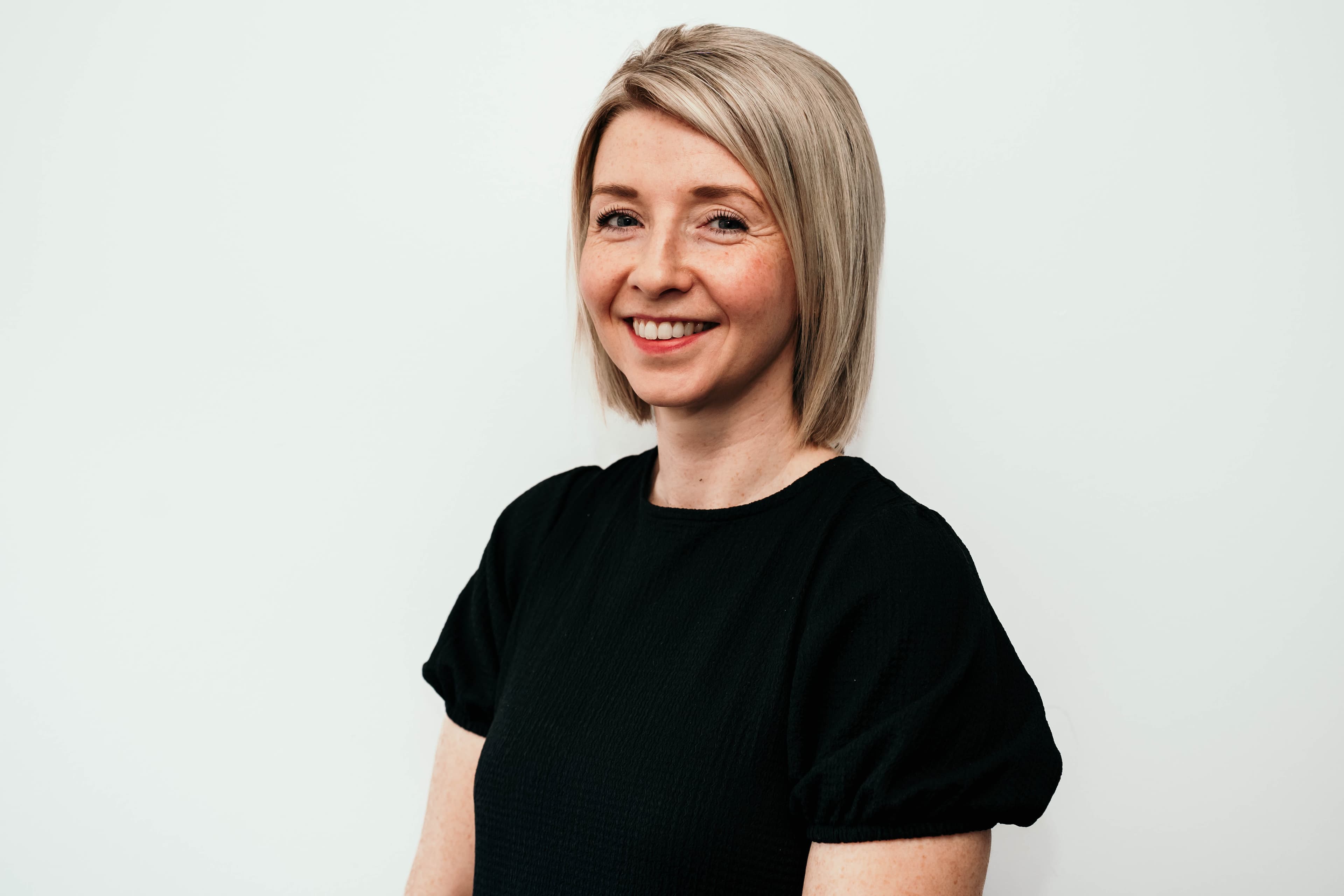 Smiling woman with short blonde hair and freckles wearing a black top against white background.