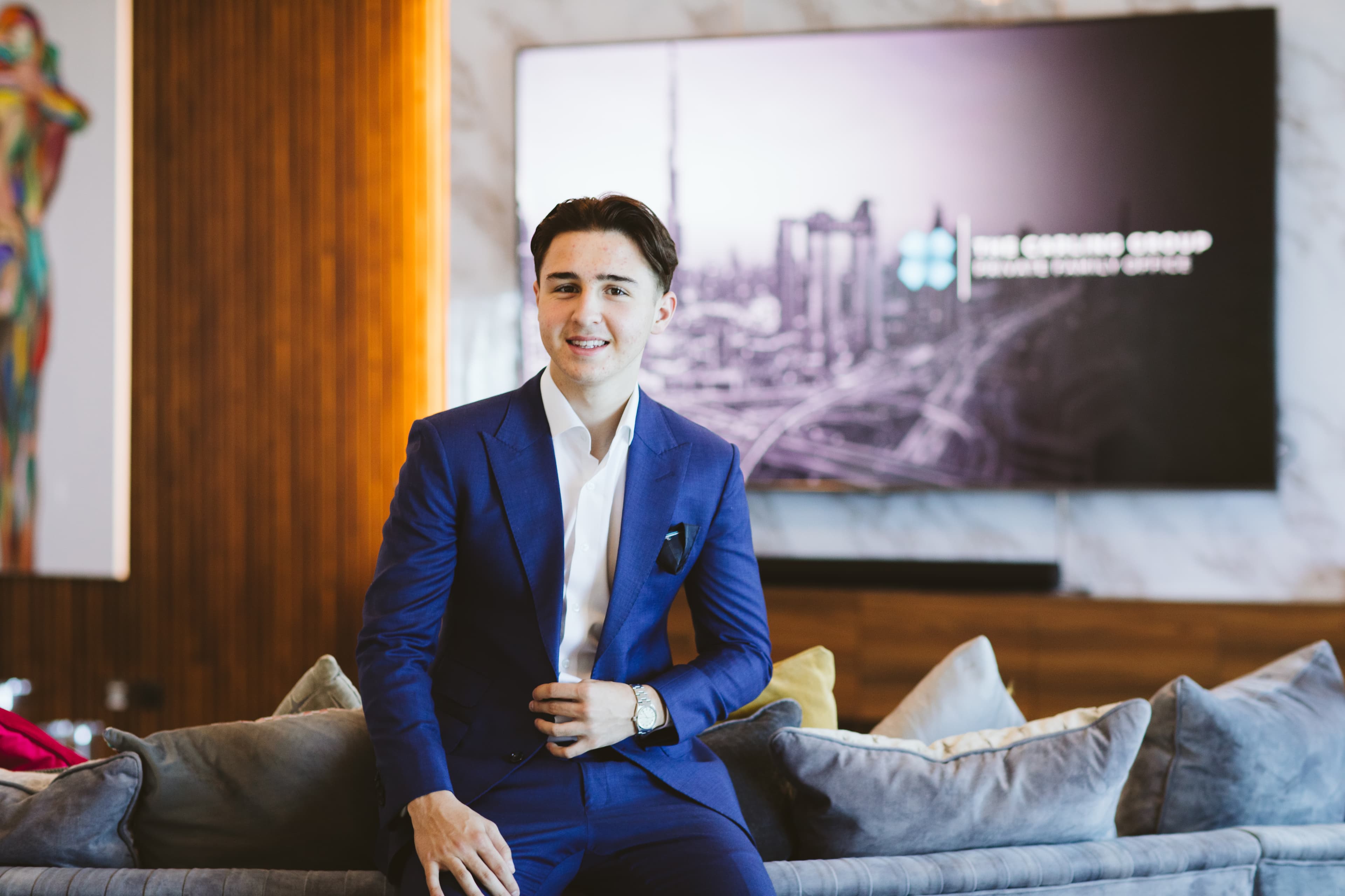 Smiling young man in a blue suit sitting on a sofa in a modern office.