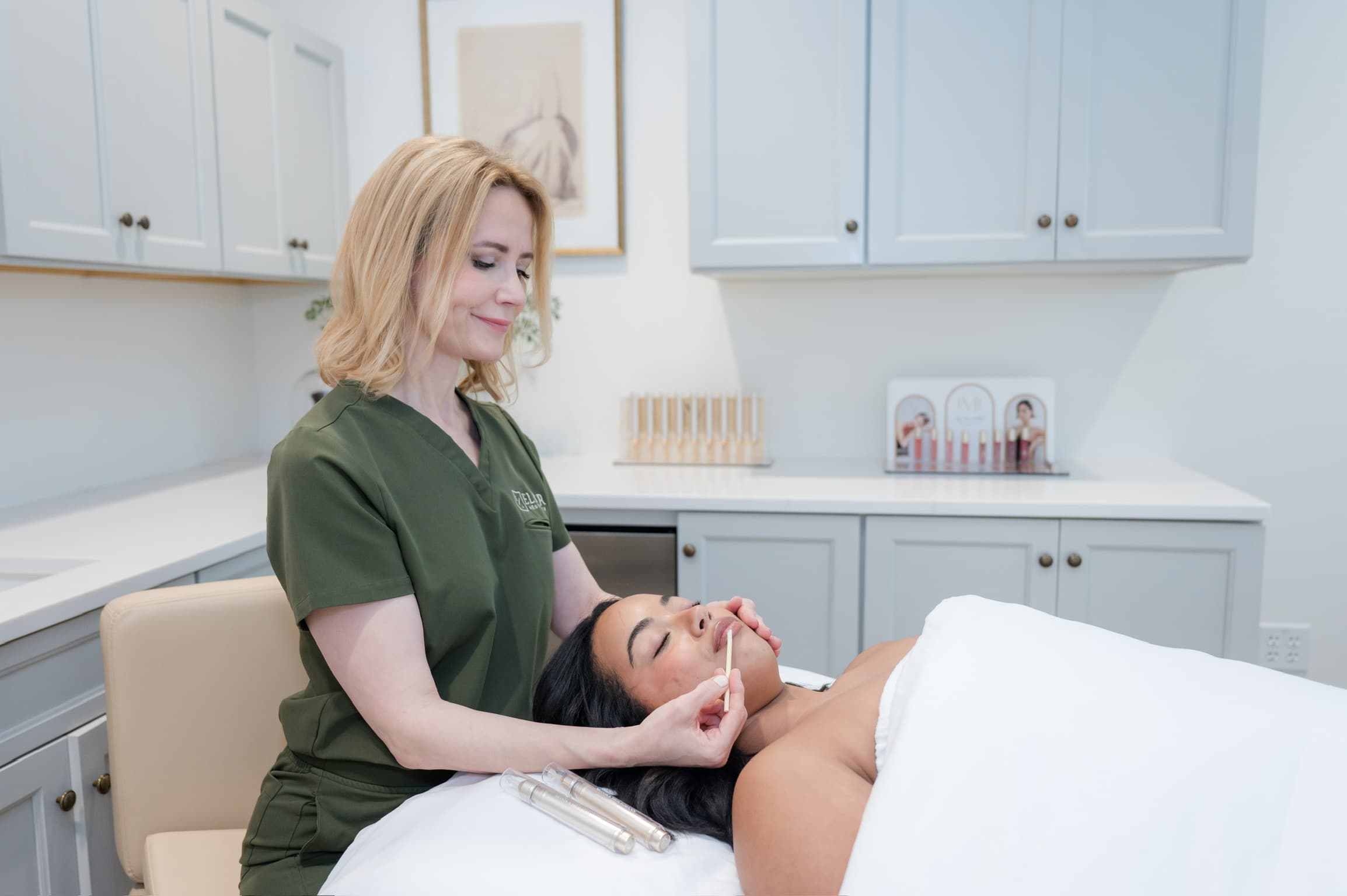 Esthetician in green scrubs applying a lip treatment to a client in a professional clinic.