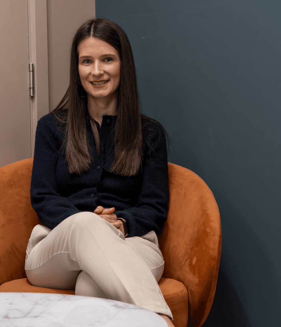 Smiling woman with long brown hair sits in an orange velvet armchair against a teal wall.